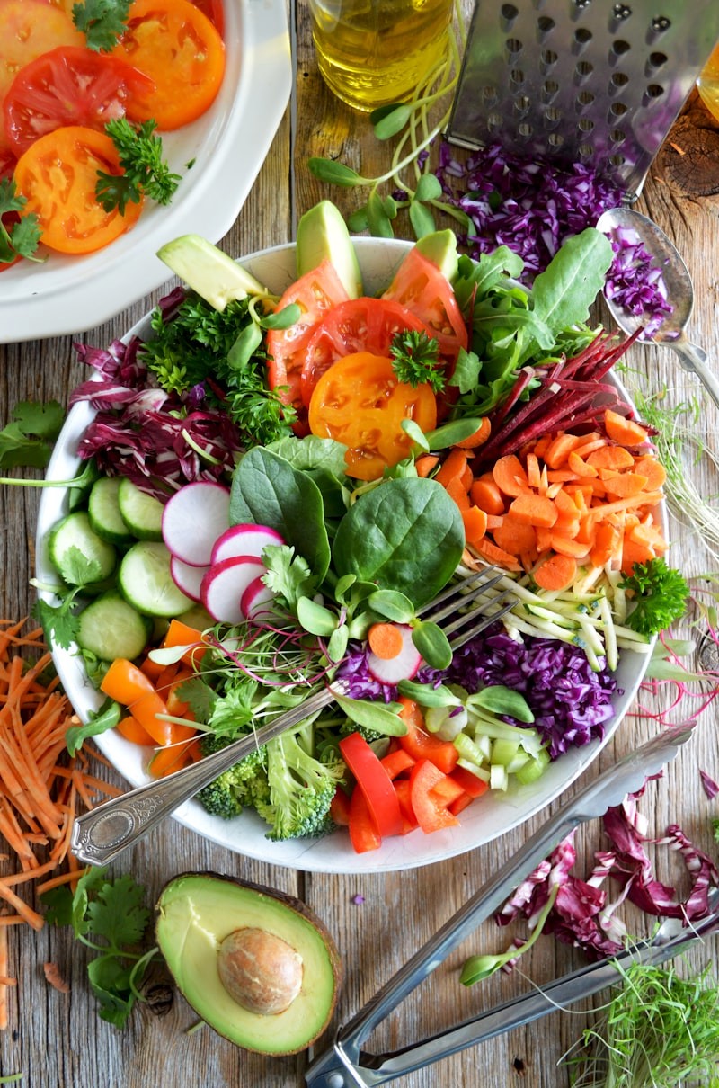 Top-down rainbow vegetable salad bowl with tomatoes, cucumber, avocado, and microgreens beside olive oil on a wooden table